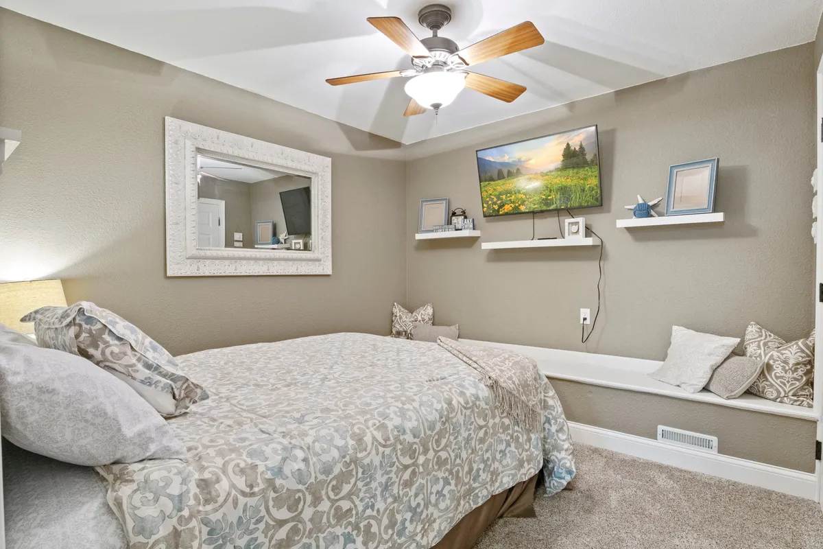 A tranquil bedroom with a patterned bed, wall-mounted television, decorative mirror, a shelf with charming items, and a cozy window seat. This space offers privacy, comfort, and the perfect nook for unwinding with a book or cup of tea.