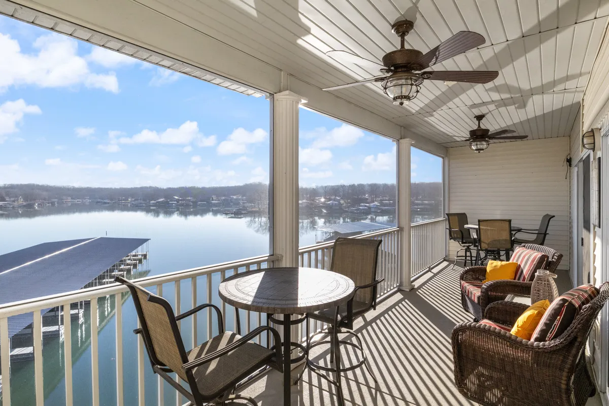The image showcases a serene, spacious outdoor seating area on a covered deck overlooking a tranquil cove of Lake of the Ozarks. Comfortable furnishings and ceiling fans invite guests to relax, while a large dining table hints at lively meals and gatherings with family or friends. The calm water, lush shoreline, and open sky set a peaceful, inviting scene perfect for both relaxation and entertaining.