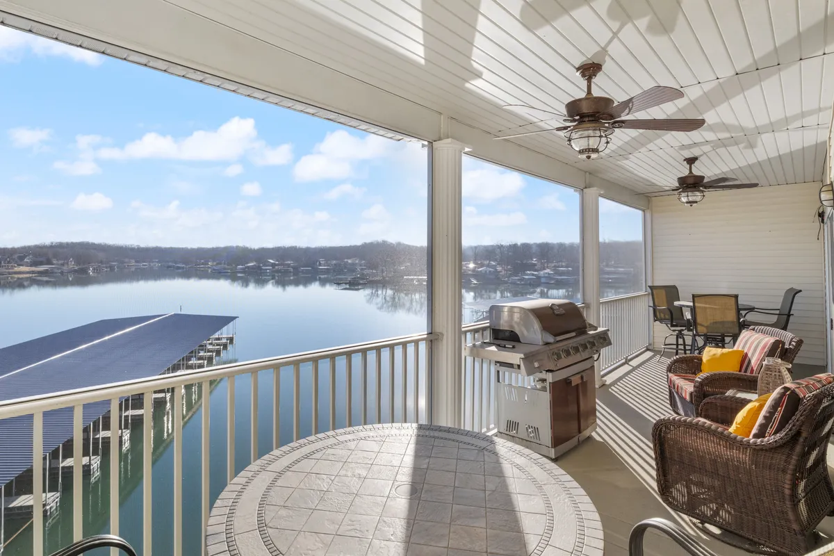 A scenic exterior shot featuring a comfortable outdoor seating area with a grill and decorative table, set against a stunning lakeside backdrop. The space suggests both daytime relaxation and sunset dinners.
