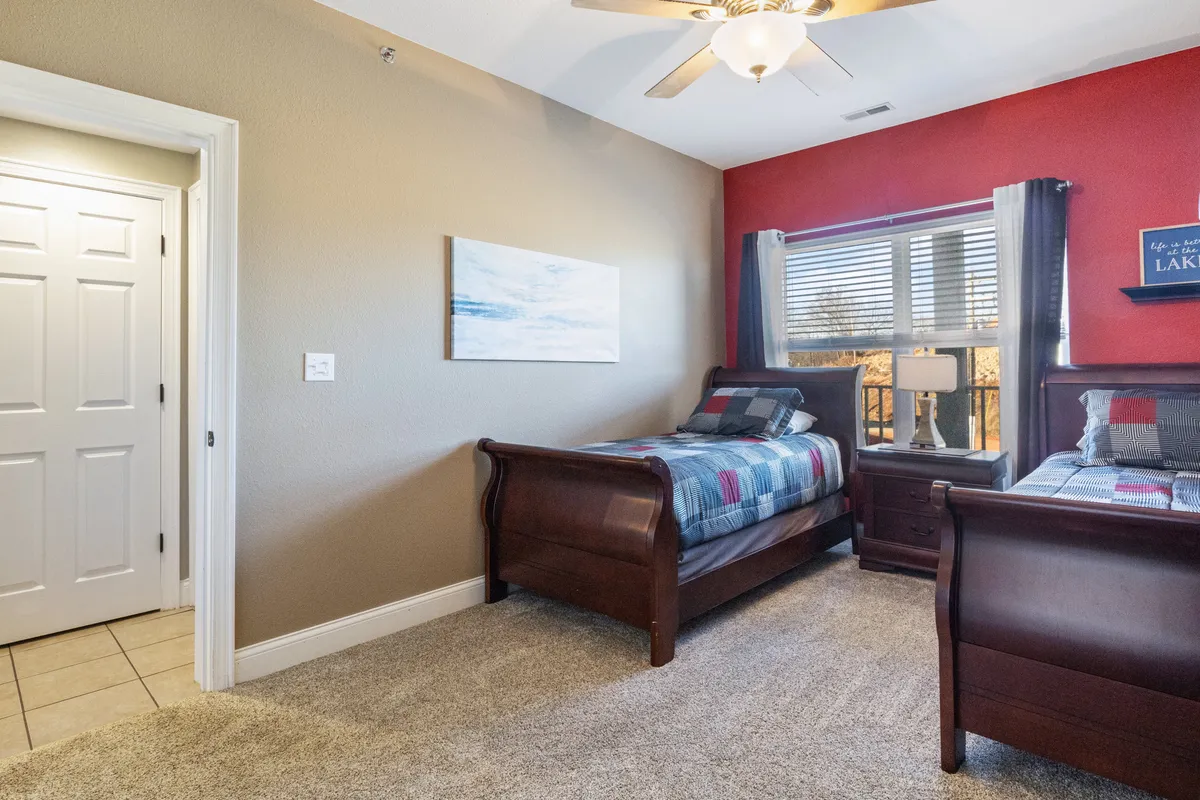A bedroom with two wooden beds dressed in colorful bedding, a window with a scenic view, a lamp on the bedside table, and framed artwork. The space feels cozy and relaxed, perfect for families or friends sharing a room.