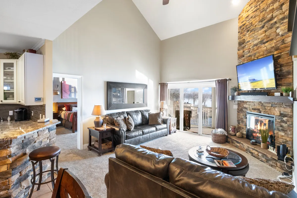 A spacious living area with a comfortable seating arrangement, a large stone fireplace, and vast windows offering clear views of the shimmering lake outside. The adjacent kitchen counter and open-plan design make it easy for groups to interact while enjoying the scenery.