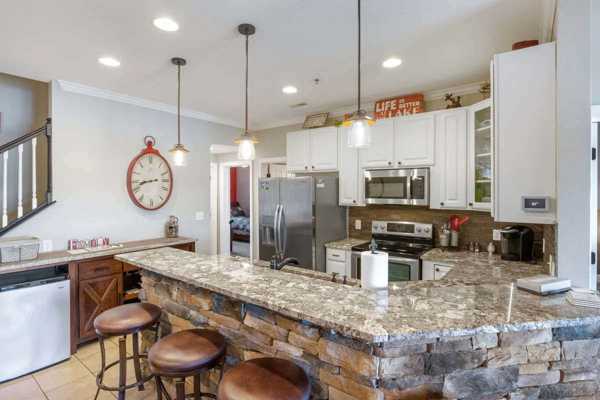 A functional and stylish kitchen space with a granite countertop, bar stools, stainless steel appliances, and pendant lighting. The wall clock and open layout enhance the inviting feel, perfect for casual meals or celebratory dinners.