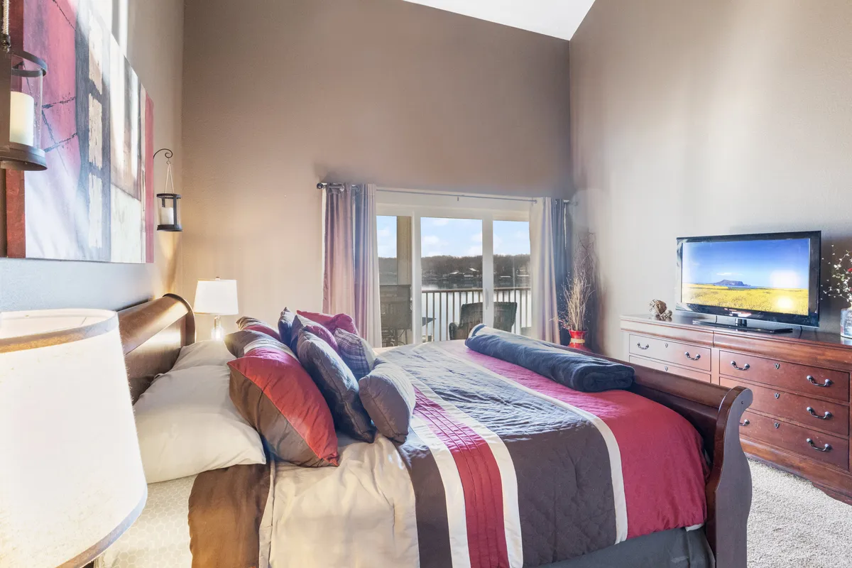 A spacious bedroom with a bed adorned in colorful bedding, large windows overlooking the water, and a wooden dresser topped with a TV. The room feels comfortable and inviting, with ample natural light and a view that draws nature indoors.