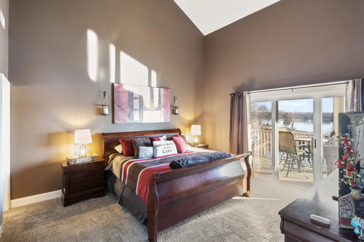 A bedroom featuring a bed with colorful, inviting bedding set against a backdrop of large windows that offer serene lake views. Decorative artwork hangs on the wall, and the room is bathed in natural light, creating a cozy and tranquil atmosphere perfect for relaxation.