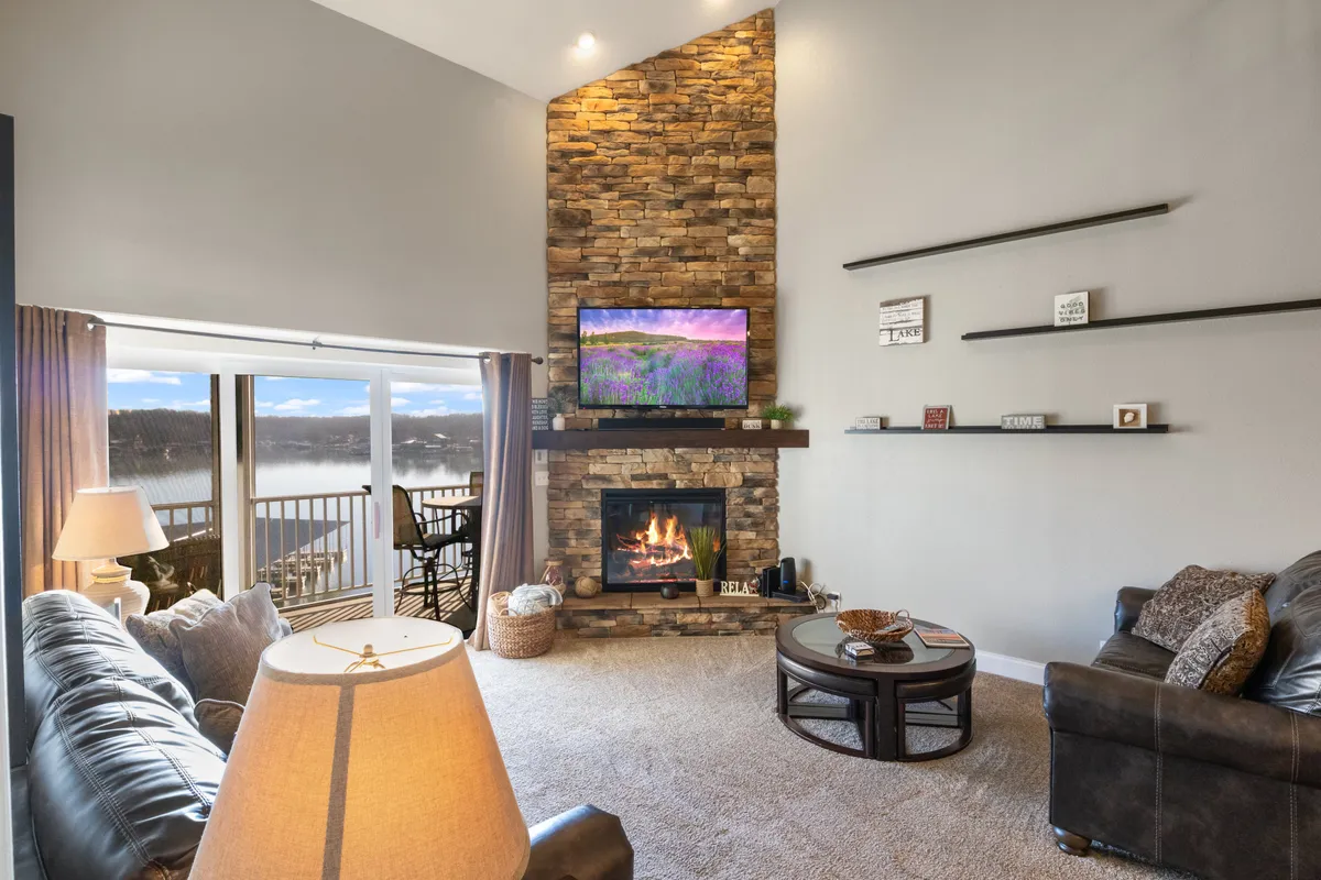 A cozy open-concept living room features comfortable seating arranged around a striking stone fireplace and a television displaying a scenic image. Expansive windows and a glass door frame beautiful lake views, seamlessly connecting the indoor gathering space to the balcony outside.