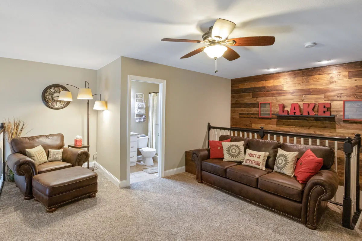 This living room features a wooden accent wall with bold 'LAKE' lettering, a comfortable leather sofa, and a reading chair with decorative pillows. The decor communicates a welcoming, home-like environment ideal for relaxing, entertaining, or spending quality family time.
