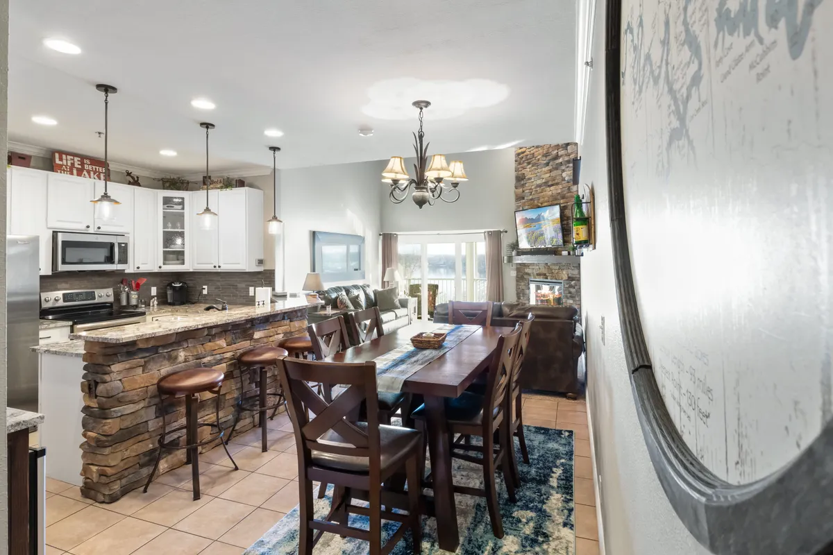 An inviting dining area showcasing a large wooden table set beneath a sparkling chandelier, adjacent to a spacious kitchen island. The open layout encourages group gatherings for lively meals and celebrations, while stone accents and warm lighting enhance the welcoming mood.